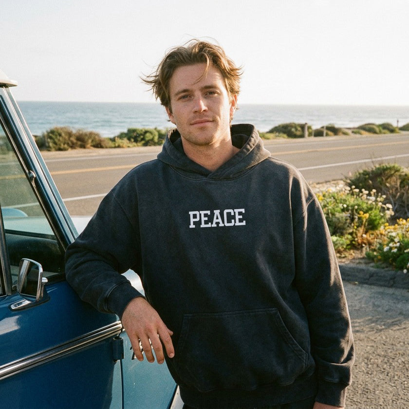 Man wearing a hoodie with 'PEACE' text, standing next to a car by the ocean.