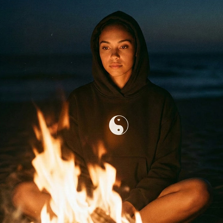 Person wearing a hoodie with a yin yang symbol sitting by a fire on a dark beach.