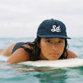 Person wearing a navy cap with 'La' logo, lying on a surfboard in the water.